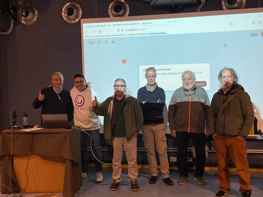 L'immagine mostra sei uomini in piedi uno accanto all'altro. Sono in piedi su un palco di fronte a una grande pantalla. I primi tre uomini sono vestiti con maglioni e camicie casuali. I tre uomini successivi indossano giacche invernali. Un microfono si trova tra i primi due uomini. L'immagine è scattata in interni, con un'illuminazione brillante.