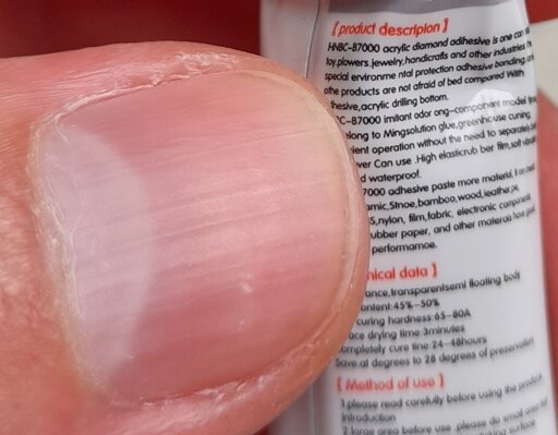 An extreme close-up of a human thumb holding a small tube of glue, which has writing on it. Comparing the size of the thumb with the size of the writing makes it clear that the print size is microscopic